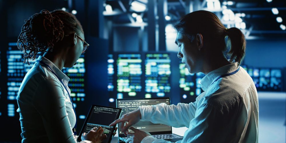 Engineering colleagues running code on tablet and laptop, updating professional data center equipment designed to accommodate server racks, networking gadgets and storage arrays